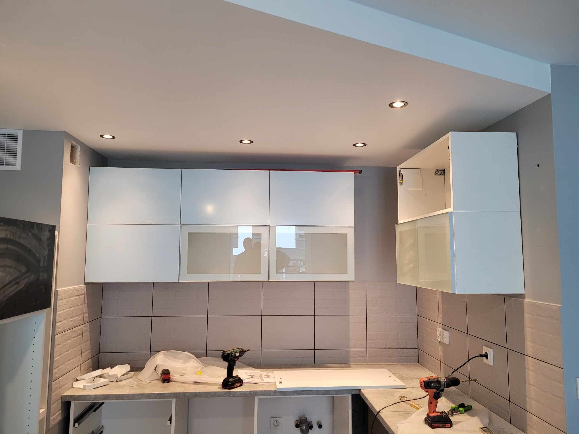 Montaż górnych szafek kuchennych z przeszklonymi frontami, widoczne wkrętarki i rozpakowane elementy, płytki ścienne w odcieniach szarości.
