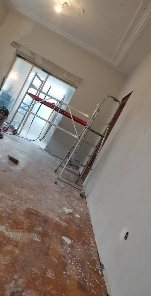 Pokój w trakcie odświeżania: białe ściany, drewniana podłoga z plamami farby, aluminiowa drabina oparta o ścianę, w tle rusztowanie przy oknie zabezpieczonym folią.