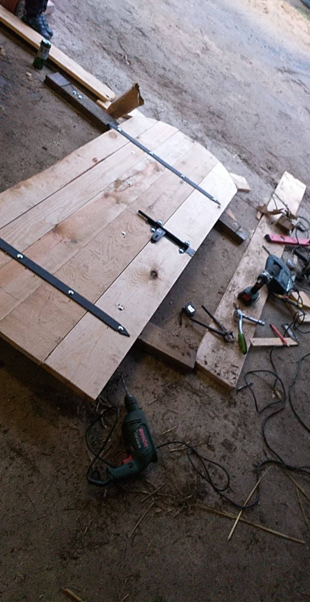 Drewniane, łukowate drzwi z czarnymi okuciami w trakcie montażu, leżące na podłodze, obok narzędzi, w tym wiertarki Bosch.