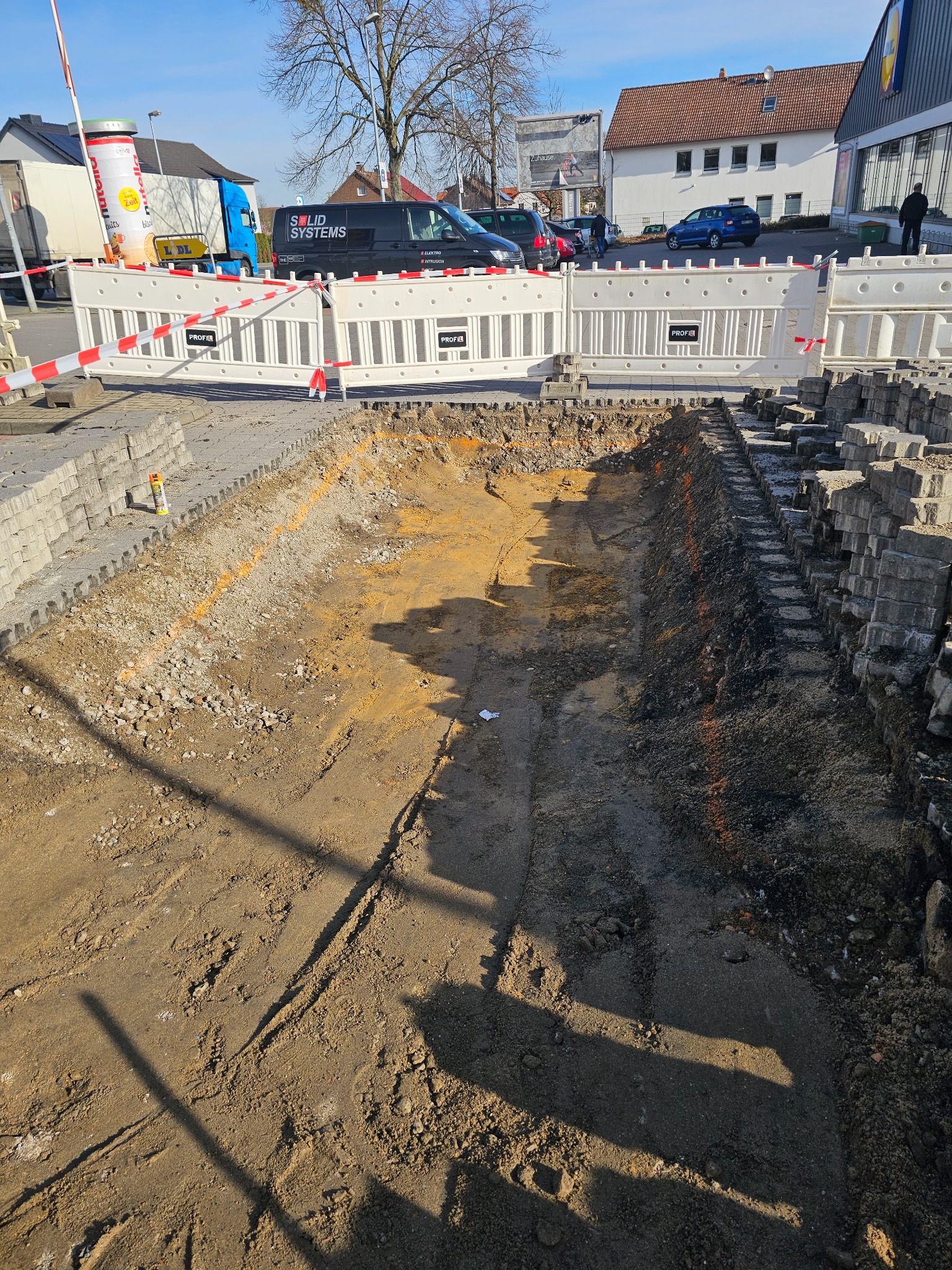 Wykop pod chodnik z kostki brukowej, widoczne warstwy ziemi i piasku, obok ułożone kostki, zabezpieczone barierkami ochronnymi, w tle budynki i samochody.