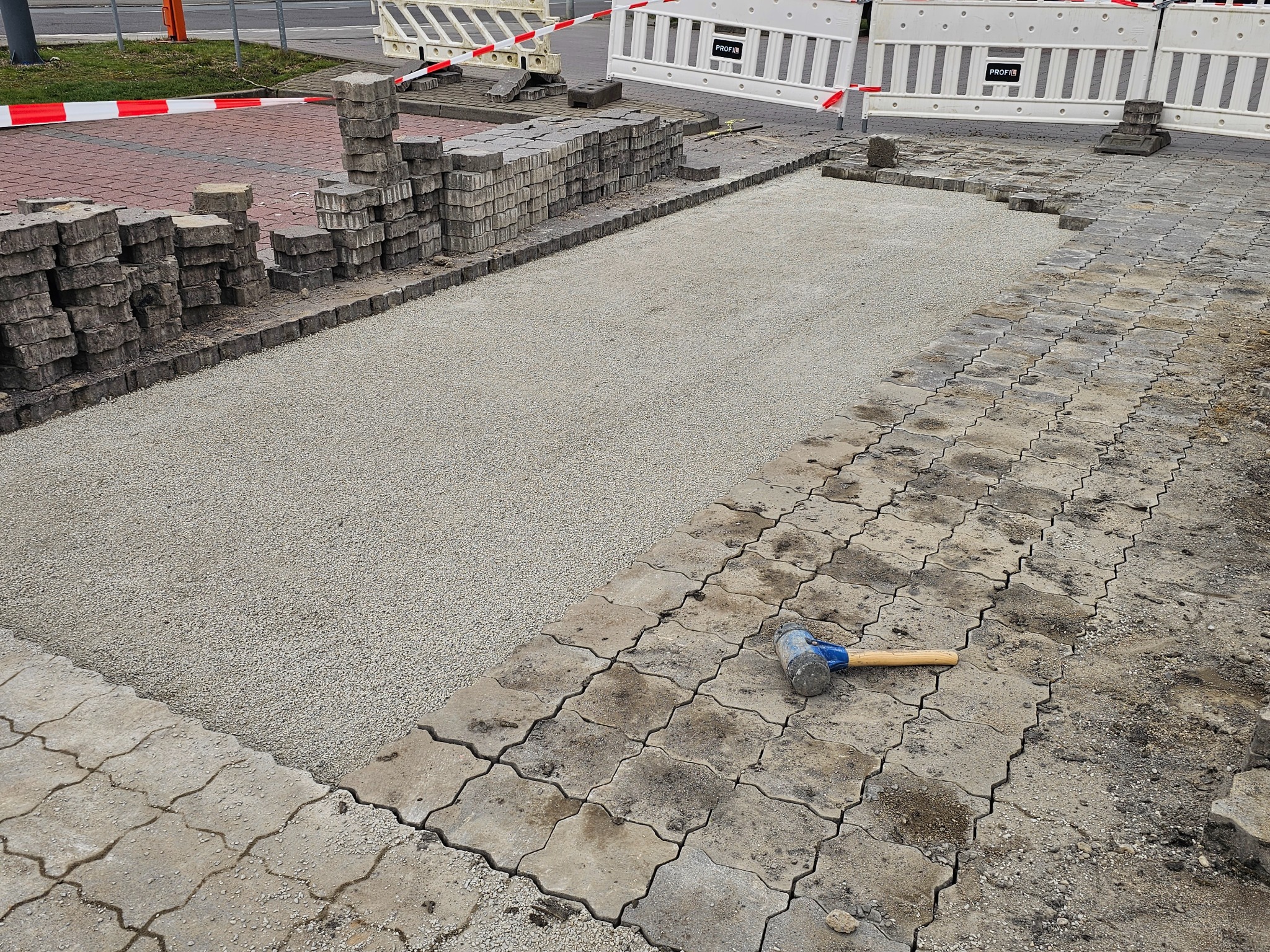 Układanie szarej kostki brukowej na podsypce piaskowej, widoczny gumowy młotek i stos kostki obok, bariery ochronne w tle.