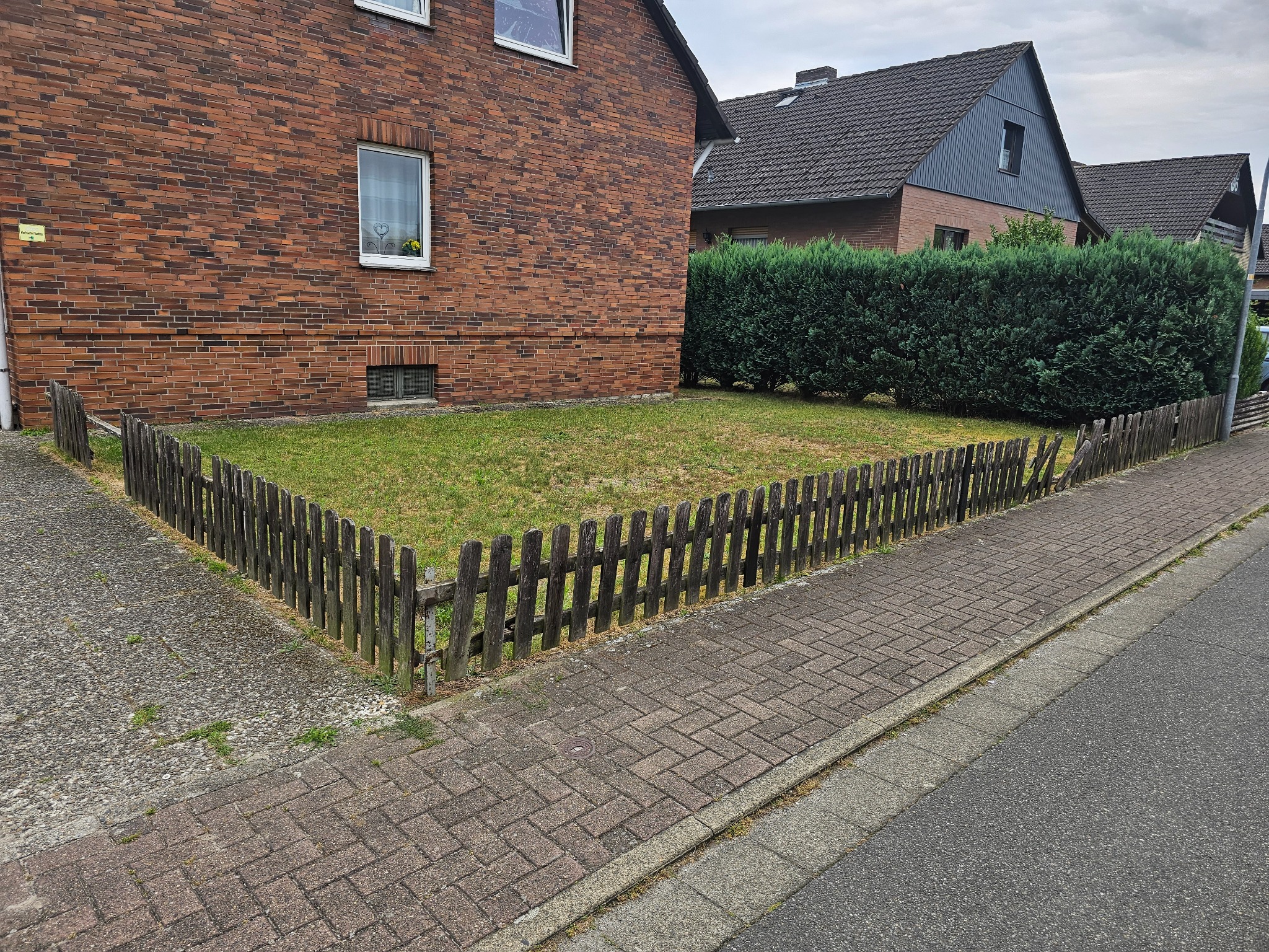Ceglany dom z drewnianym płotem otaczającym trawnik, widok z chodnika z kostki brukowej.