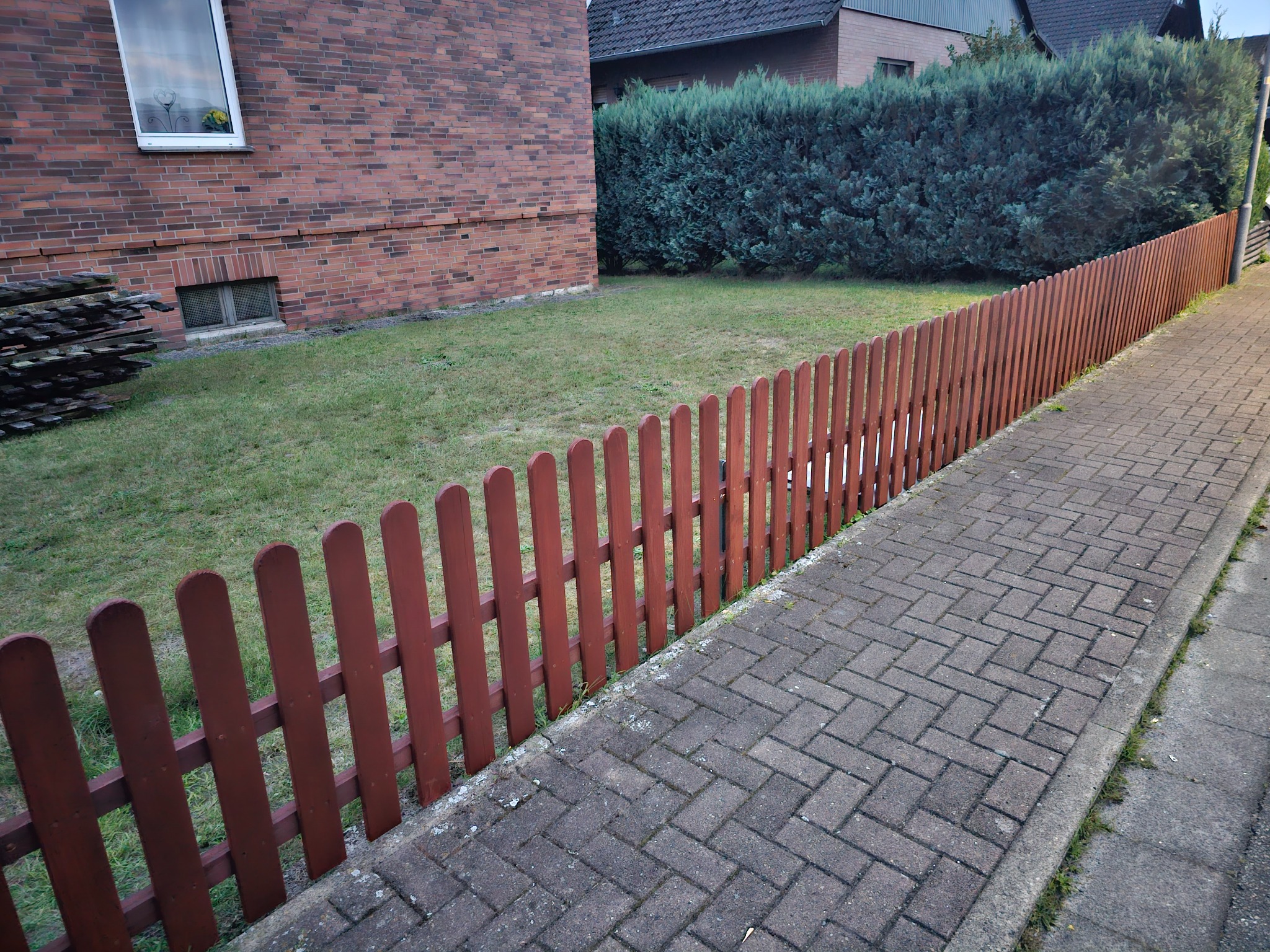 Świeżo pomalowany, niski, drewniany płot w kolorze ceglastym oddziela trawnik od chodnika z kostki brukowej przed domem z czerwonej cegły i zielonym żywopłotem.