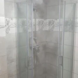 Narożna kabina prysznicowa z chromowaną armaturą i szklanymi drzwiami, otoczona szarymi płytkami z dekoracyjnym pasem z motywem kwiatowym.