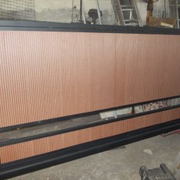 Metaloplastyka , konstrukcje stalowe , spawanie, ślusarka - Nowoczesna brama przesuwna z ciemną metalową ramą i pionowymi panelami w kolorze rdzawego brązu, widoczna w warsztacie przed montażem.