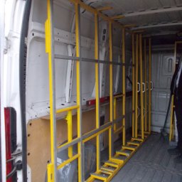 Metaloplastyka , konstrukcje stalowe , spawanie, ślusarka - Wnętrze białego samochodu dostawczego z zamontowanym jaskrawożółtym, metalowym regałem transportowym o stopniowanej podstawie, przeznaczonym do przewozu płaskich elementów.
