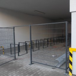Metaloplastyka , konstrukcje stalowe , spawanie, ślusarka - Szara, ażurowa brama z otwartym skrzydłem, zabezpieczająca wjazd do zadaszonego parkingu rowerowego z rzędami stojaków. Widoczne żółto-czarne słupki ochronne i kostka brukowa.
