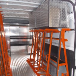 Metaloplastyka , konstrukcje stalowe , spawanie, ślusarka - Wnętrze dostawczego samochodu z pomarańczowymi, metalowymi stojakami typu A-frame, ryflowanymi aluminiowymi panelami na ścianach i podłodze oraz szafkami.