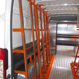 Metaloplastyka , konstrukcje stalowe , spawanie, ślusarka - Wnętrze białego furgonu z rzędem pomarańczowych, metalowych stojaków z czarnymi gumowymi zabezpieczeniami, do transportu płaskich elementów. Podłoga i ściana wyłożone ryflowaną blachą.