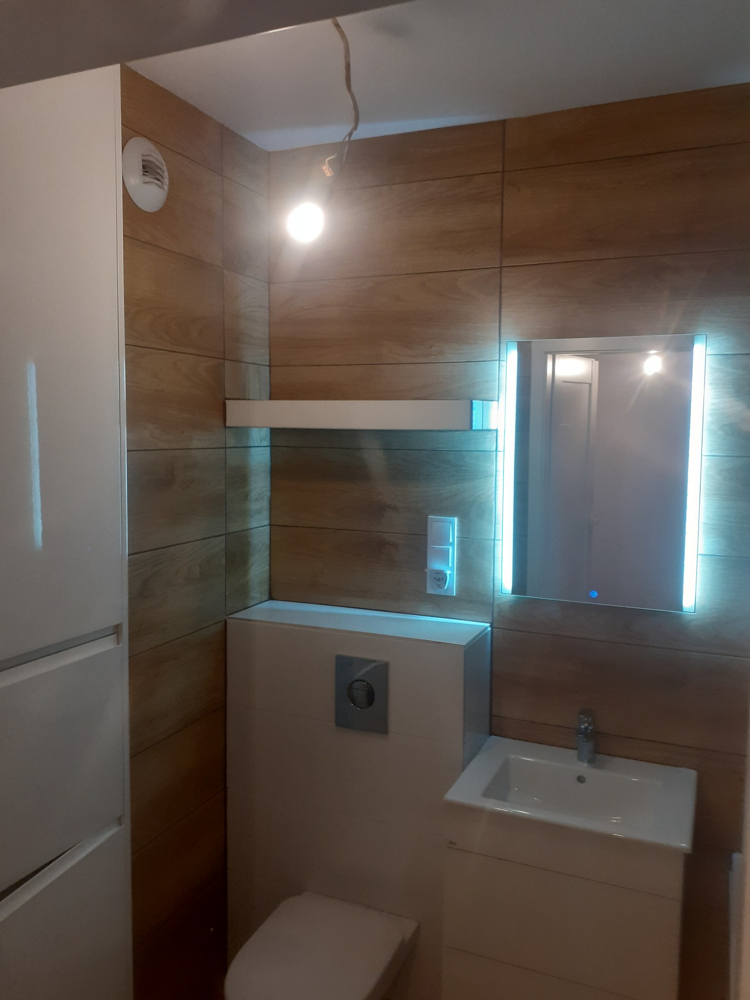 Nowoczesna łazienka z drewnopodobnymi płytkami, podświetlanym lustrem, białą szafką i wiszącą toaletą, minimalistyczny styl.