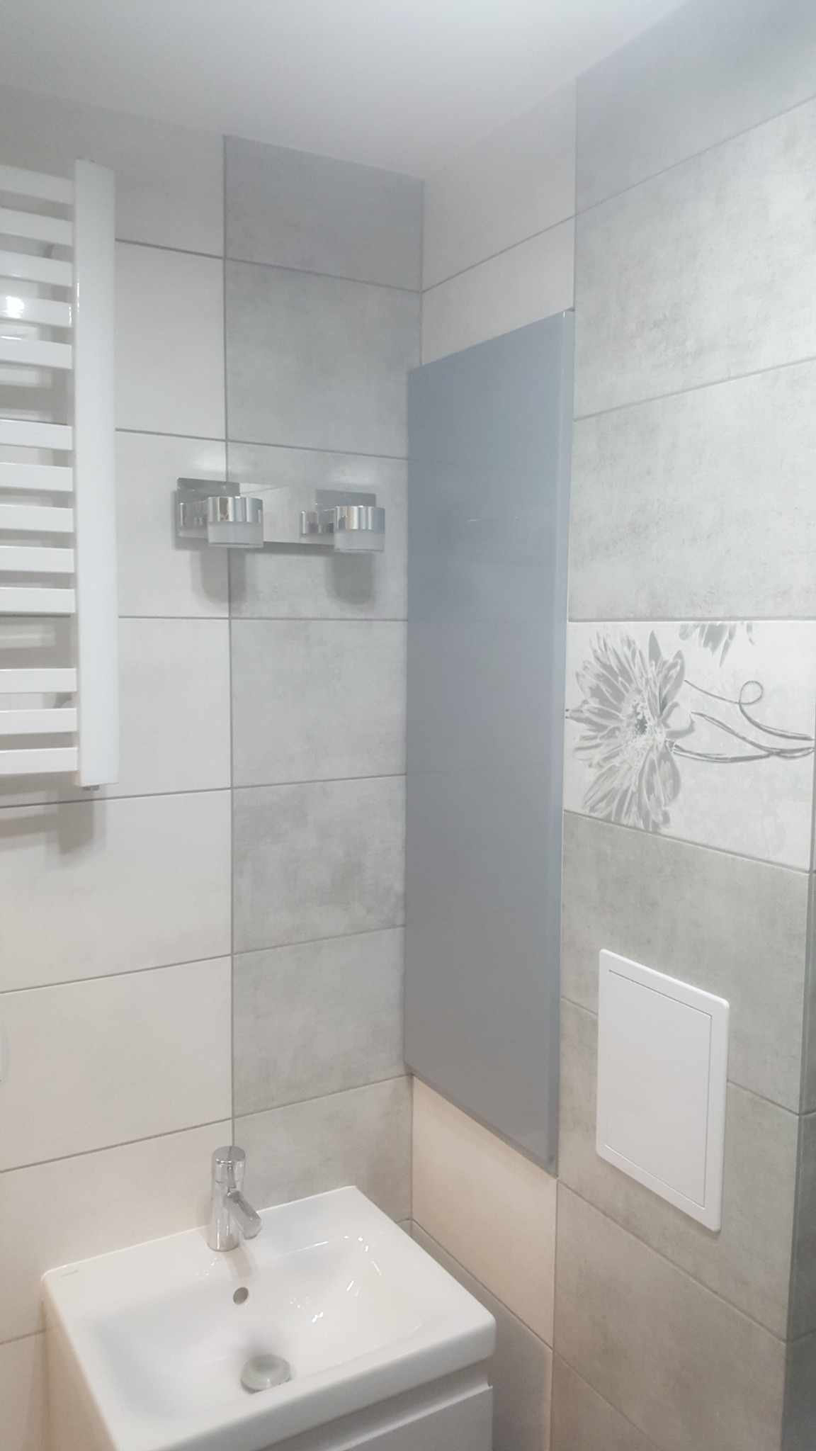 Wykończona łazienka w szaro-białych odcieniach z kwadratowymi płytkami, umywalką nablatową z chromowaną baterią, grzejnikiem drabinkowym i dekoracyjną płytką z motywem kwiatowym.