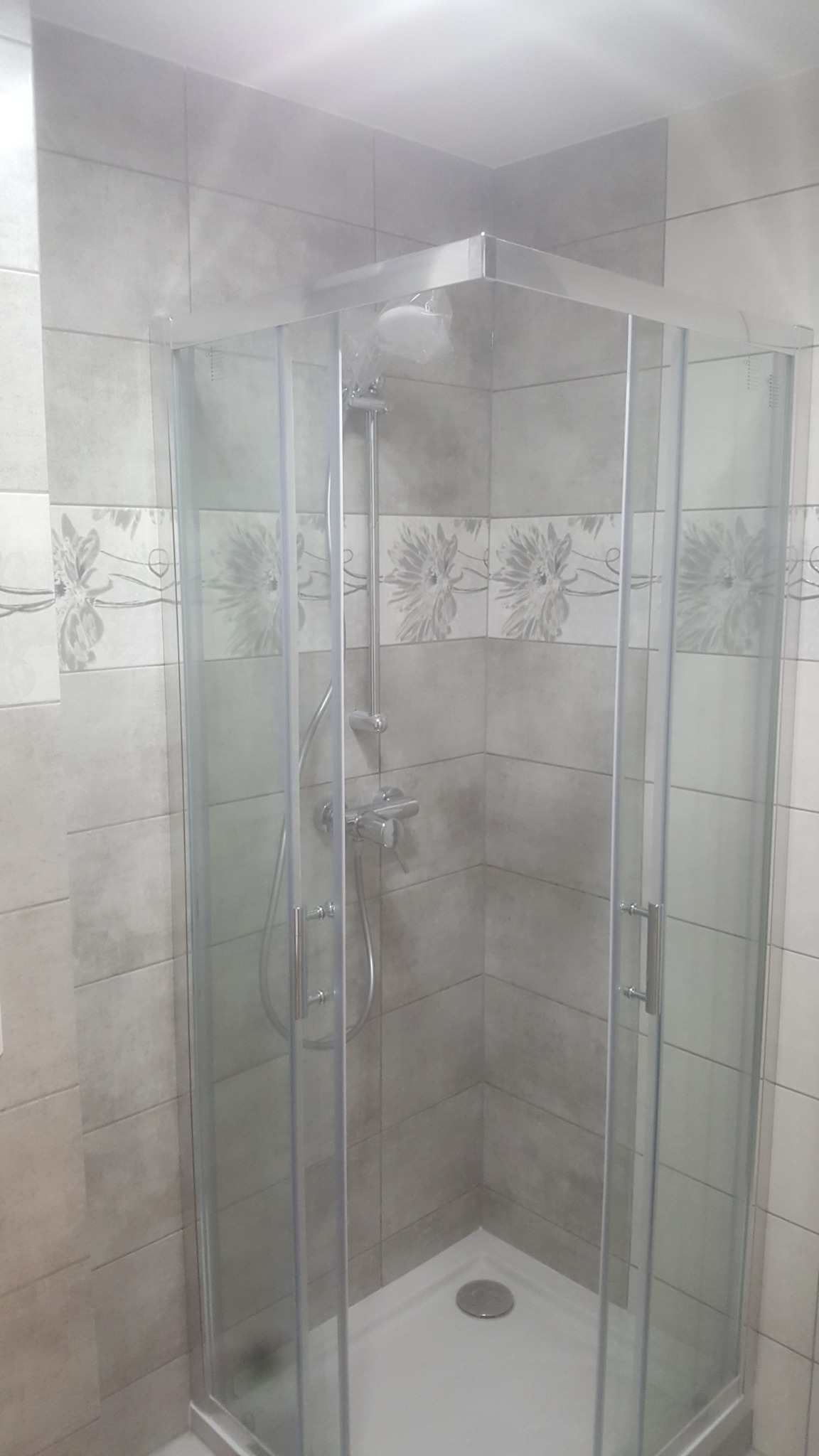 Narożna kabina prysznicowa z chromowaną armaturą i szklanymi drzwiami, otoczona szarymi płytkami z dekoracyjnym pasem z motywem kwiatowym.