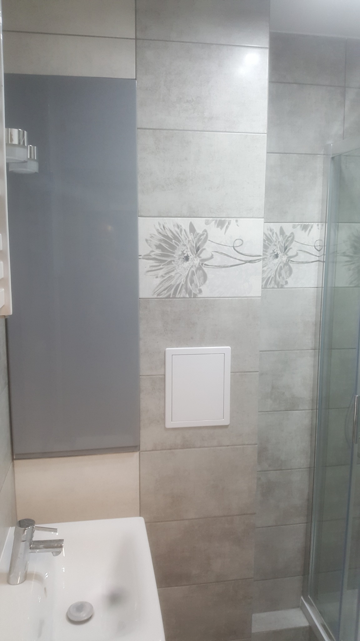 Umywalka z chromowaną baterią i szara szafka w łazience wykończonej płytkami w odcieniach szarości z dekoracyjnym pasem z motywem kwiatowym, widoczny fragment kabiny prysznicowej.