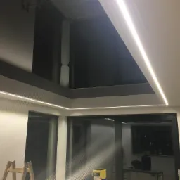Nowoczesny sufit z czarnym, lustrzanym wykończeniem i liniowym oświetleniem LED wzdłuż krawędzi. Widoczny fragment drabiny i sprzęt budowlany sugerują prace wykończeniowe.