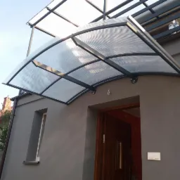 Zadaszenie nad wejściem do domu z poliwęglanu i metalową konstrukcją, widok z dołu, szara elewacja budynku.