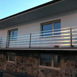 Nowoczesna balustrada balkonowa z poziomych, jasnych profili zamontowana na balkonie domu z elewacją z kamienia, widoczna osoba na balkonie.