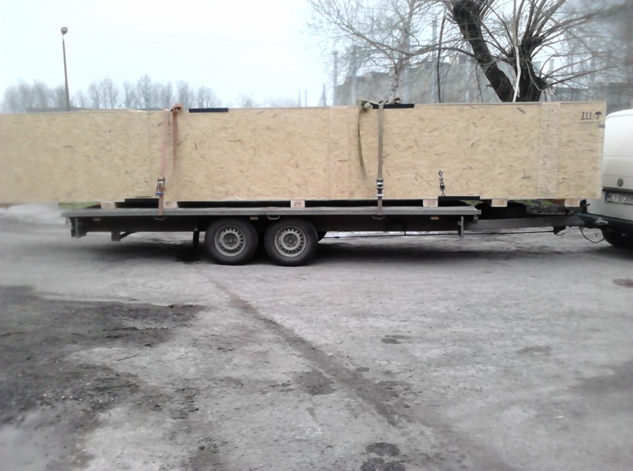 Duża skrzynia z płyty OSB zabezpieczona pasami transportowymi na przyczepie dwuosiowej, widok z boku na szarym betonowym podłożu w pochmurny dzień.