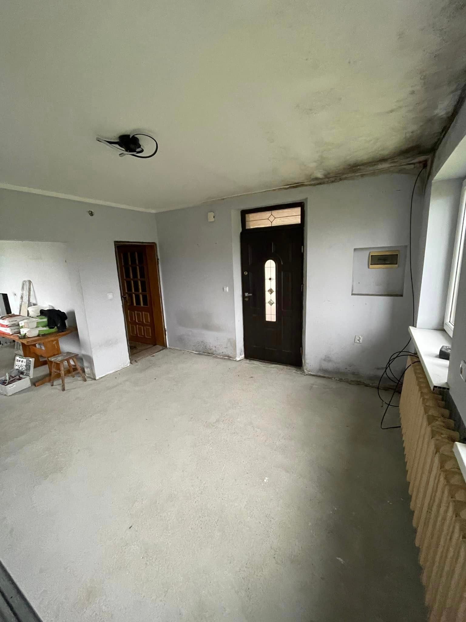 Pokój w trakcie remontu: surowe ściany z widocznymi śladami wilgoci i grzyba na suficie, przygotowany do dalszych prac wykończeniowych, widoczny stary grzejnik i prowizoryczne oświetlenie.