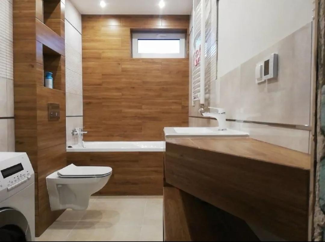 Nowoczesna łazienka z drewnopodobnymi płytkami na ścianach, podwieszana toaleta i umywalka z blatem z ciemnego drewna, oświetlenie punktowe.