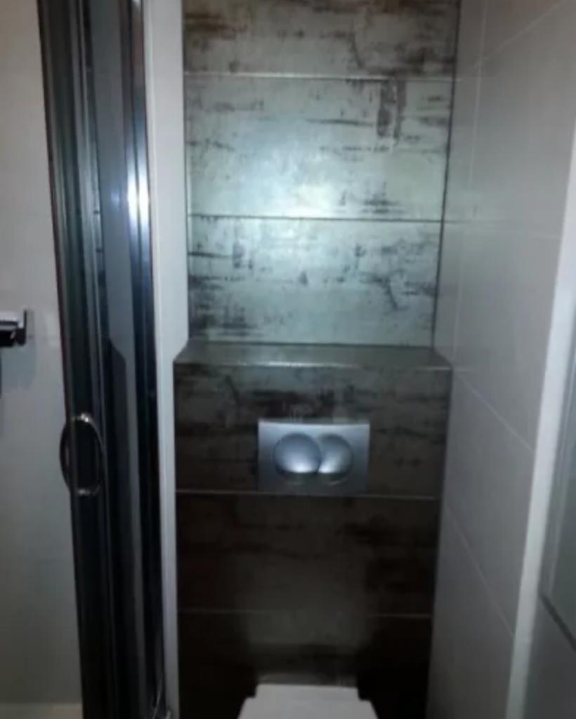 Nowoczesna łazienka z kabiną prysznicową i podwieszanym WC z płytkami imitującymi drewno na ścianie za toaletą oraz chromowanym przyciskiem spłukującym.