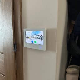 MIKOLI.pl Piotr Kowalski - Zamontowany, biały panel wideodomofonu Hikvision na ścianie wewnątrz domu, obok drewnianych drzwi. Na ekranie widok z kamery na zewnątrz budynku.