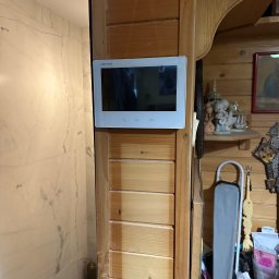 MIKOLI.pl Piotr Kowalski - Zamontowany wideodomofon Hikvision na drewnianej ścianie wewnątrz domu, obok wejścia do łazienki z marmurowymi płytkami. Widoczne elementy wystroju wnętrza.