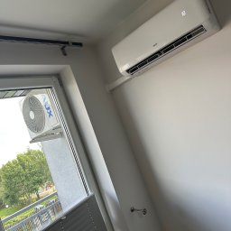 MIKOLI.pl Piotr Kowalski - Nowoczesny, biały klimatyzator ścienny AUX z cyfrowym wyświetlaczem '18' zamontowany w jasnym pomieszczeniu. Za oknem widoczna jednostka zewnętrzna i zielony krajobraz z placem zabaw.