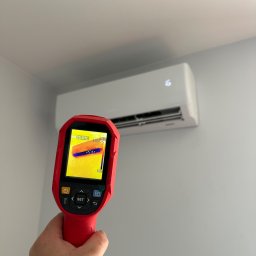 MIKOLI.pl Piotr Kowalski - Ręka trzyma czerwoną kamerę termowizyjną, pokazującą obraz termiczny 25.5°C, skierowaną na biały klimatyzator z cyfrowym wyświetlaczem '26' na jasnoszarej ścianie.