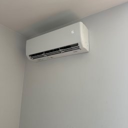 MIKOLI.pl Piotr Kowalski - Biały klimatyzator typu split z cyfrowym wyświetlaczem '16', zamontowany wysoko w narożniku jasnoszarego pokoju, z widocznym wylotem powietrza.