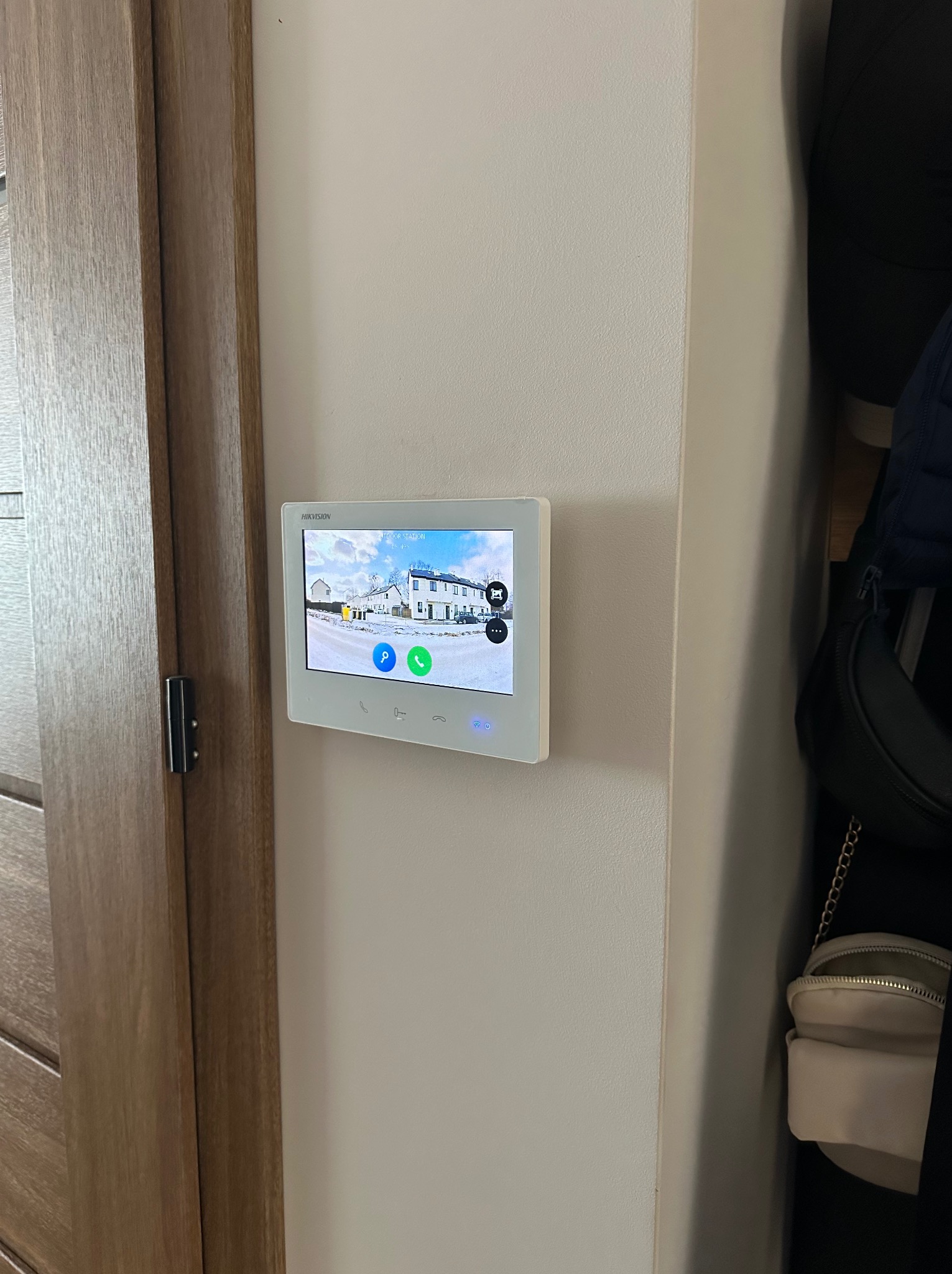 Zamontowany, biały panel wideodomofonu Hikvision na ścianie wewnątrz domu, obok drewnianych drzwi. Na ekranie widok z kamery na zewnątrz budynku.