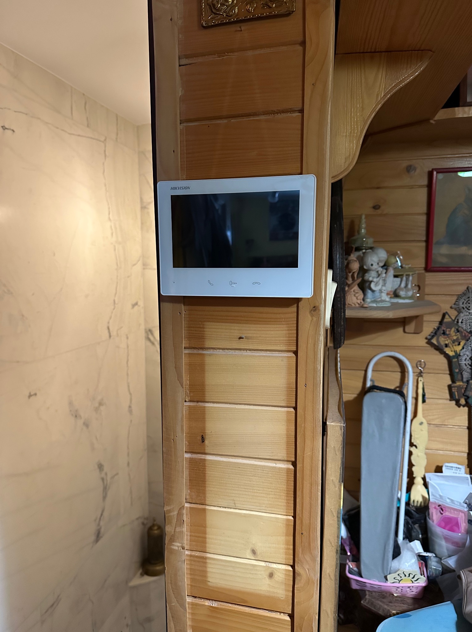Zamontowany wideodomofon Hikvision na drewnianej ścianie wewnątrz domu, obok wejścia do łazienki z marmurowymi płytkami. Widoczne elementy wystroju wnętrza.