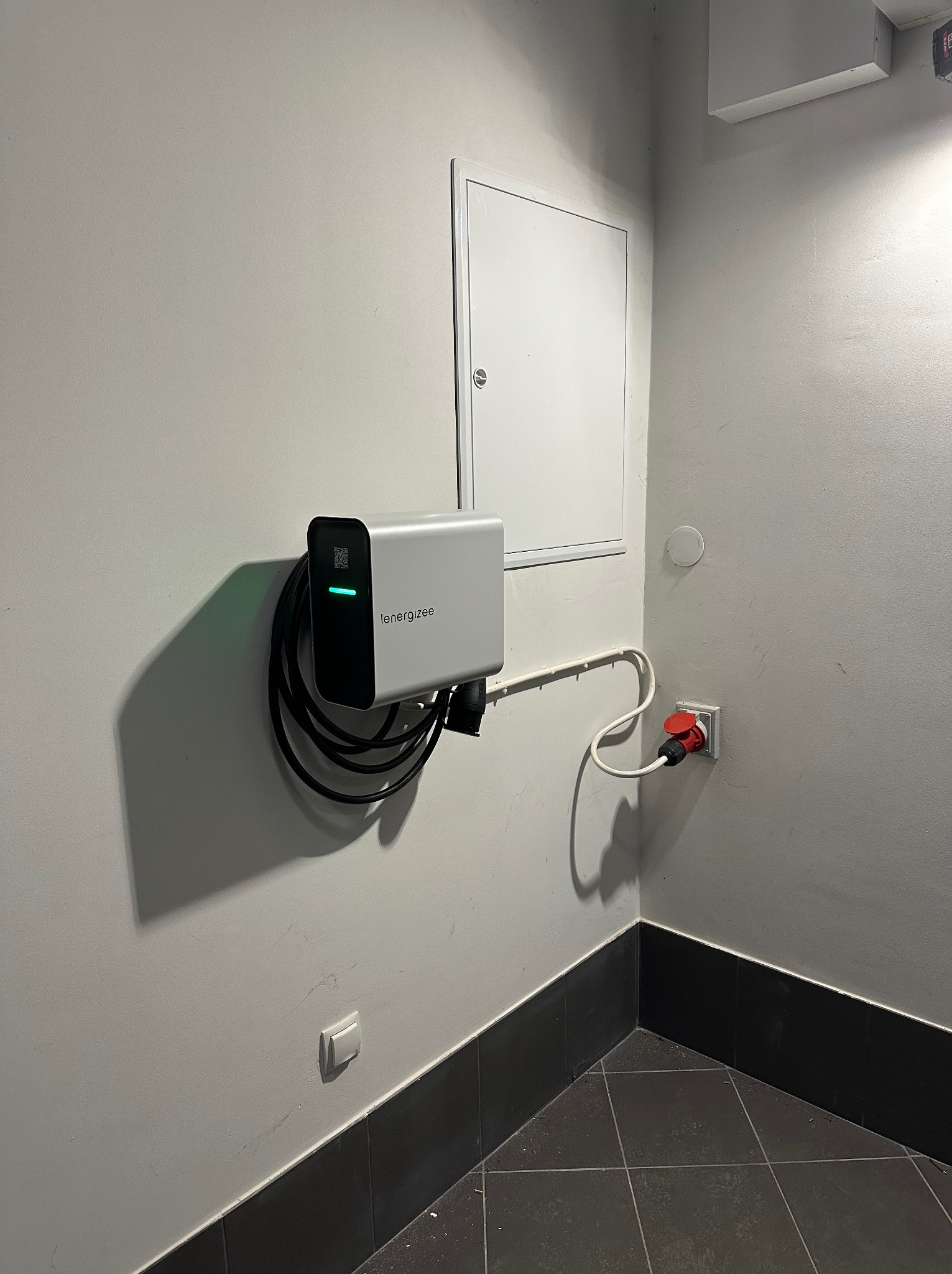 Zamontowana na ścianie ładowarka do samochodu elektrycznego marki Energizee z podłączonym kablem i wtyczką do gniazdka. Widoczna skrzynka elektryczna i listwa przypodłogowa.