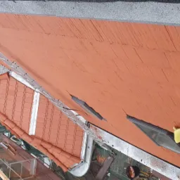 Widok z góry na płaski dach pokryty pomarańczową papą oraz fragment dachu spadzistego z czerwonej dachówki ceramicznej z zamontowaną rynną, drabina oparta o dach, w tle zabudowa miejska.