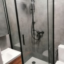 Andrii Korytchuk GLOBAL-REM - Nowoczesna kabina prysznicowa z czarnymi profilami, szare płytki ścienne, biała umywalka z drewnianą szafką i toaleta w tle.