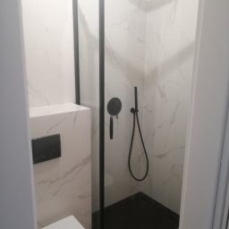 Andrii Korytchuk GLOBAL-REM - Nowoczesna kabina prysznicowa narożna z czarnymi profilami, deszczownicą i baterią, białe płytki imitujące marmur, wisząca toaleta z czarnym przyciskiem spłukiwania.