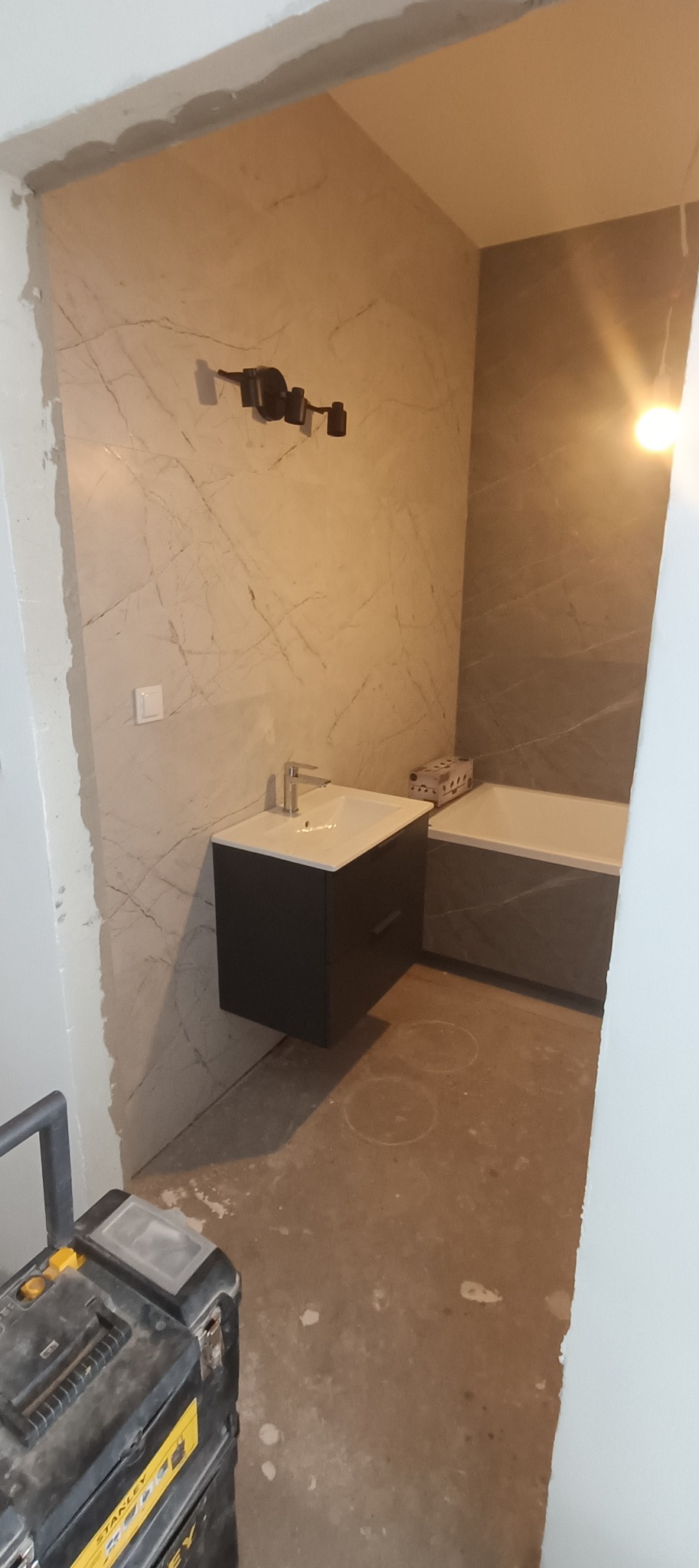 Niedokończona łazienka z marmurowymi płytkami, wiszącą szafką umywalkową i wanną, widoczny czarny reflektor sufitowy i skrzynka narzędziowa Stanley na podłodze.