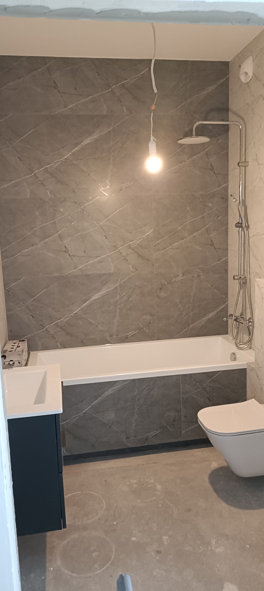 Wnętrze łazienki w trakcie remontu: ściany wyłożone szarymi płytkami imitującymi marmur, biała wanna, umywalka z szafką w kolorze granatowym, toaleta, widoczne ślady prac na podłodze, surowa...