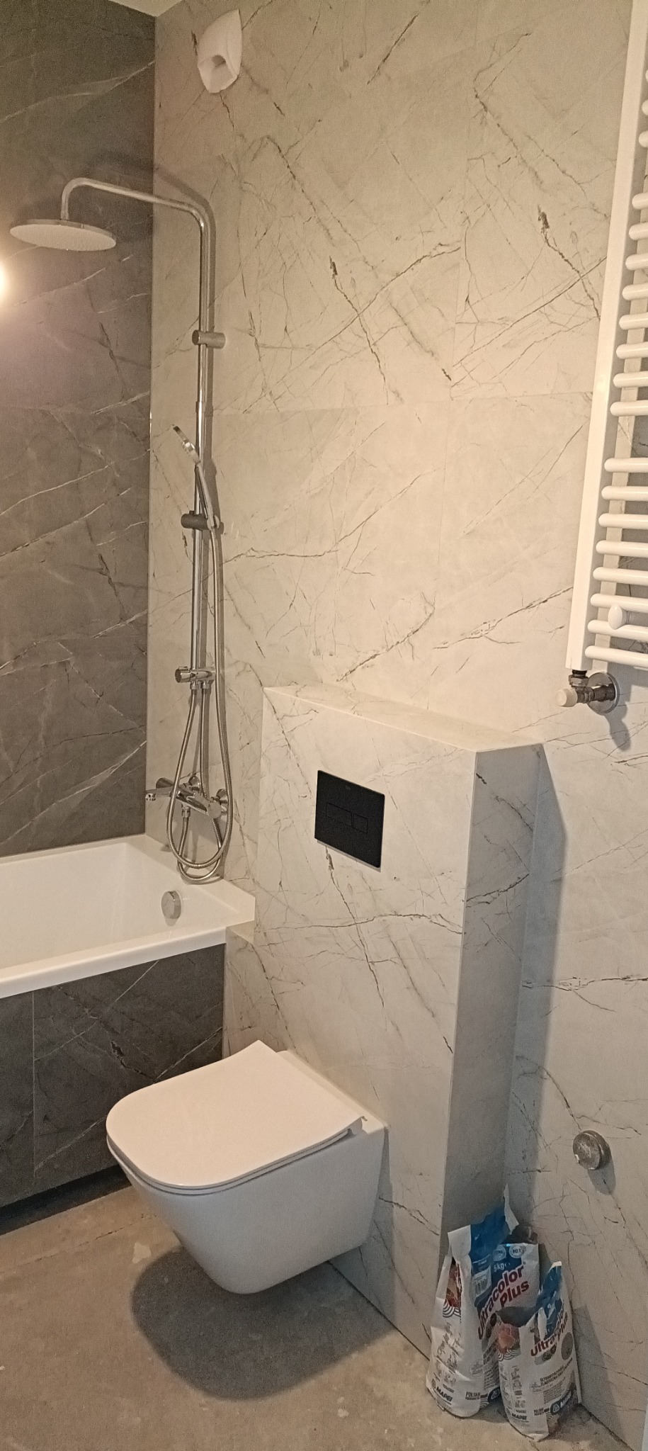 Nowoczesna łazienka z marmurowymi płytkami, wiszącą toaletą, grzejnikiem drabinkowym i workami zaprawy klejowej na podłodze.