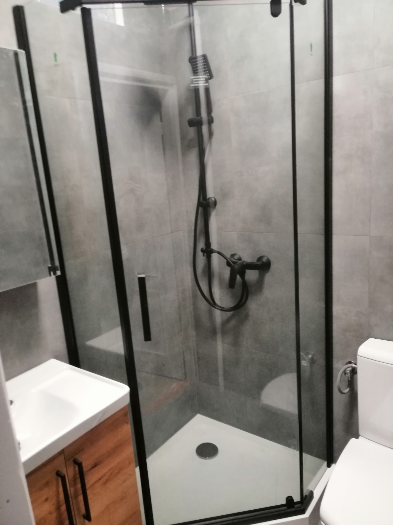 Nowoczesna kabina prysznicowa z czarnymi profilami, szare płytki ścienne, biała umywalka z drewnianą szafką i toaleta w tle.