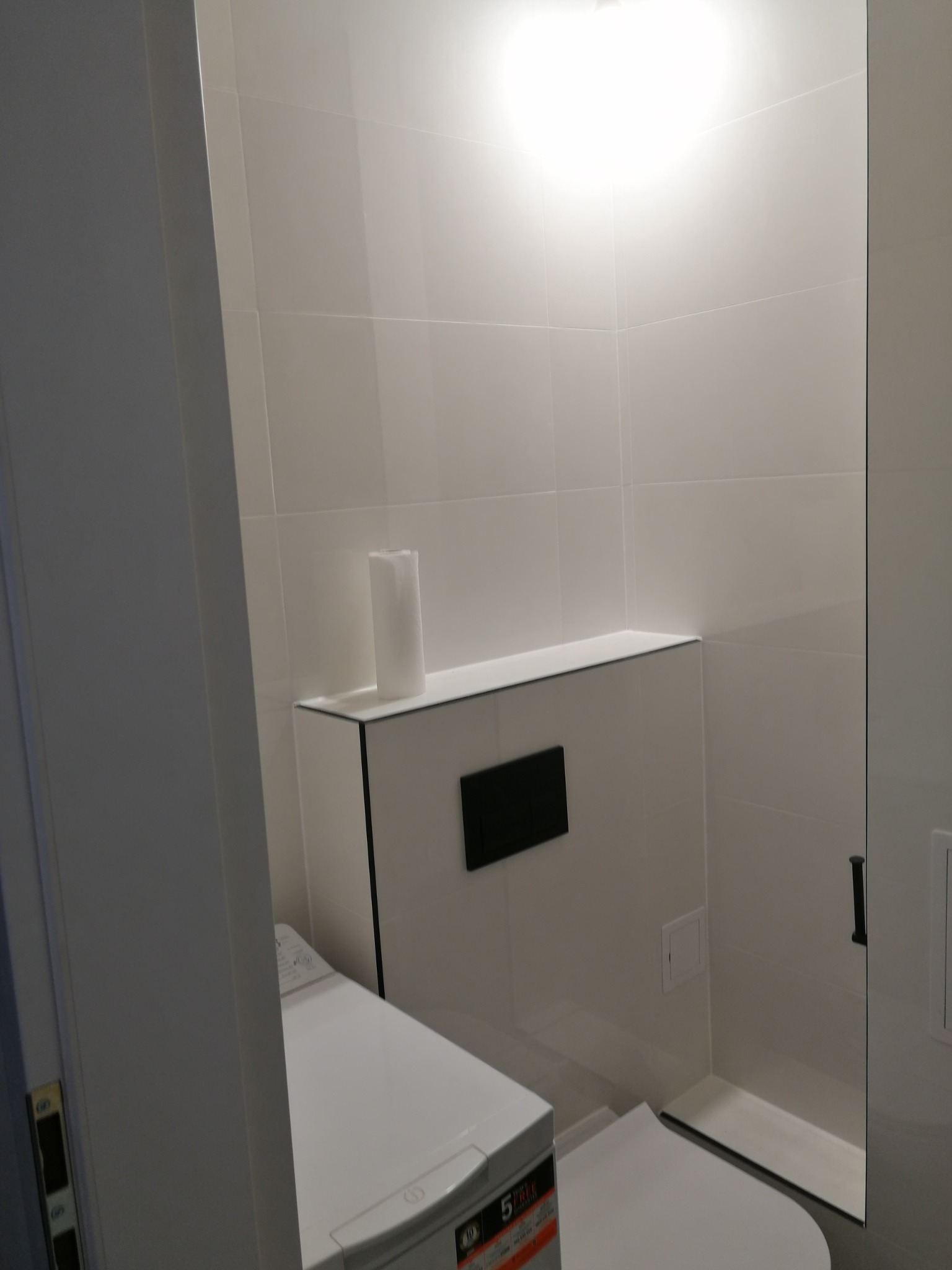 Nowoczesna łazienka z białymi płytkami, podwieszaną toaletą i pralką, minimalistyczny design, oświetlenie sufitowe.