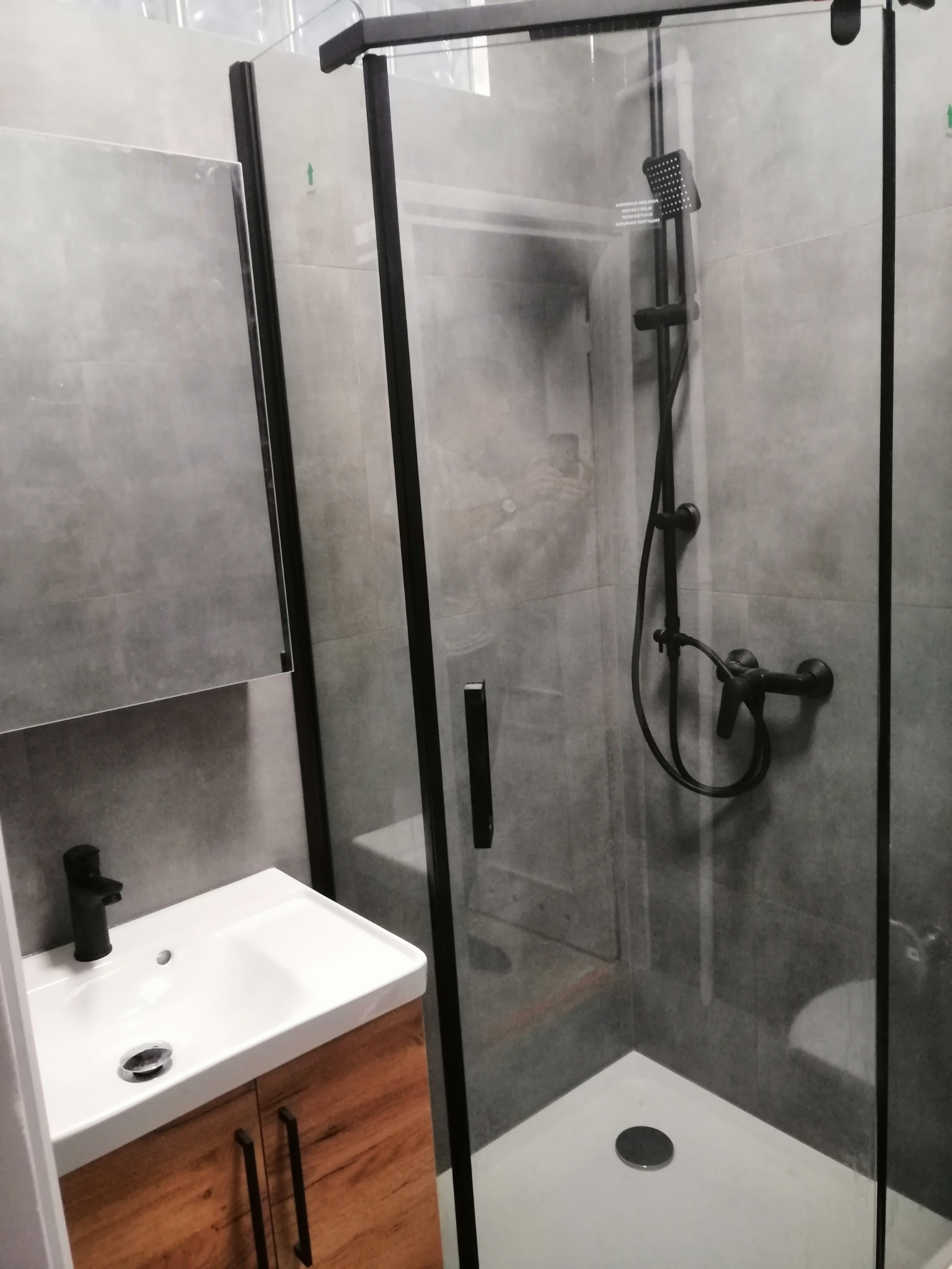 Nowoczesna łazienka z kabiną prysznicową z czarnymi profilami, umywalką nablatową z szafką z drewnopodobnym frontem i szarą glazurą.