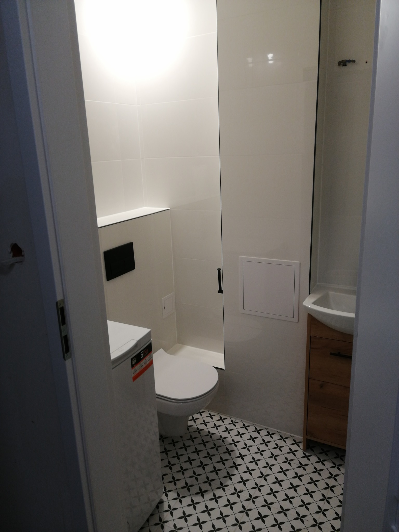 Nowoczesna łazienka z białą armaturą, umywalką w drewnianej szafce i wzorzystą podłogą z płytek ceramicznych, widziana zza uchylonych drzwi.