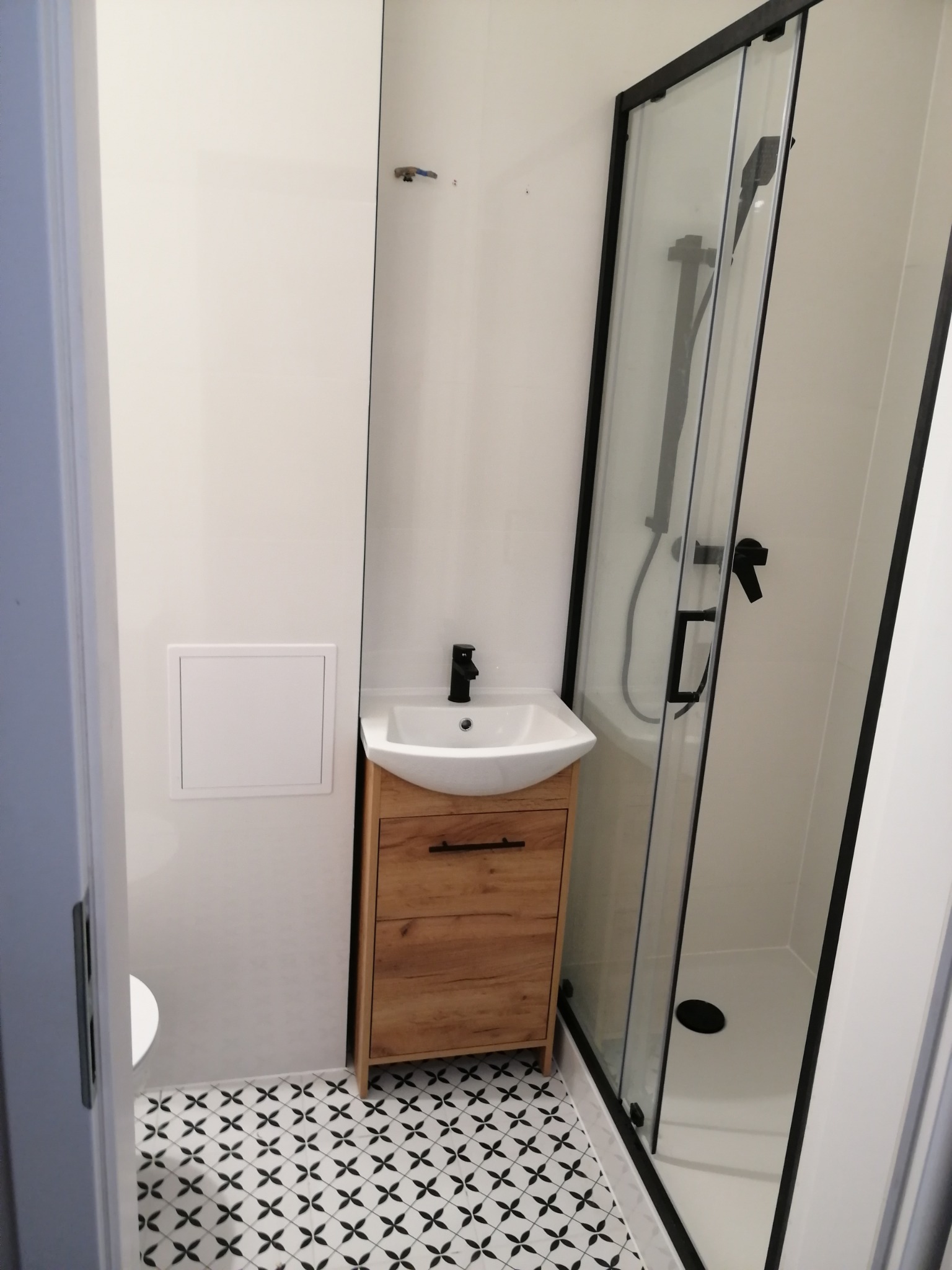 Nowoczesna łazienka z kabiną prysznicową z czarną ramą, szafką pod umywalką z drewnopodobnym frontem i czarną armaturą oraz biało-czarną wzorzystą podłogą.