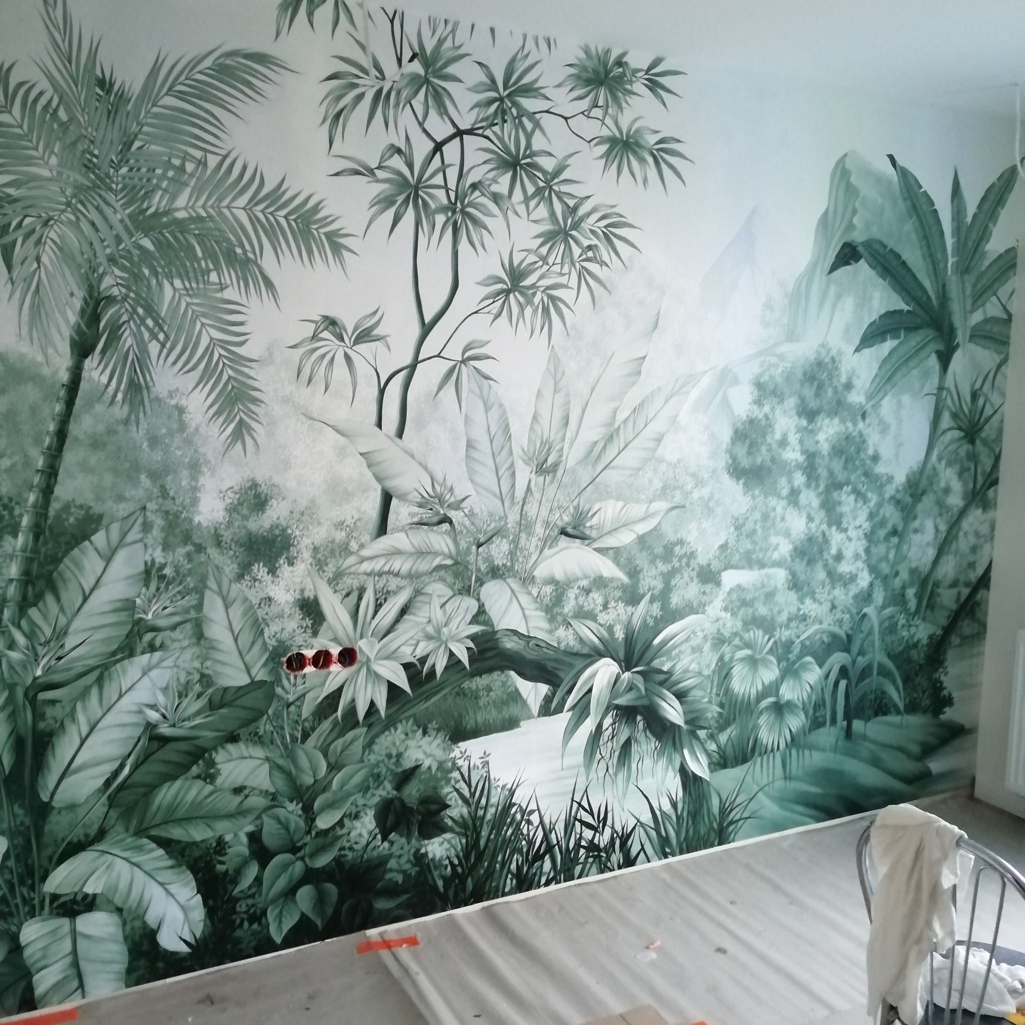 Ściana z ręcznie malowanym motywem dżungli w odcieniach szarości, widoczne puszki elektryczne i zabezpieczona podłoga, sugerująca świeżo zakończone prace.