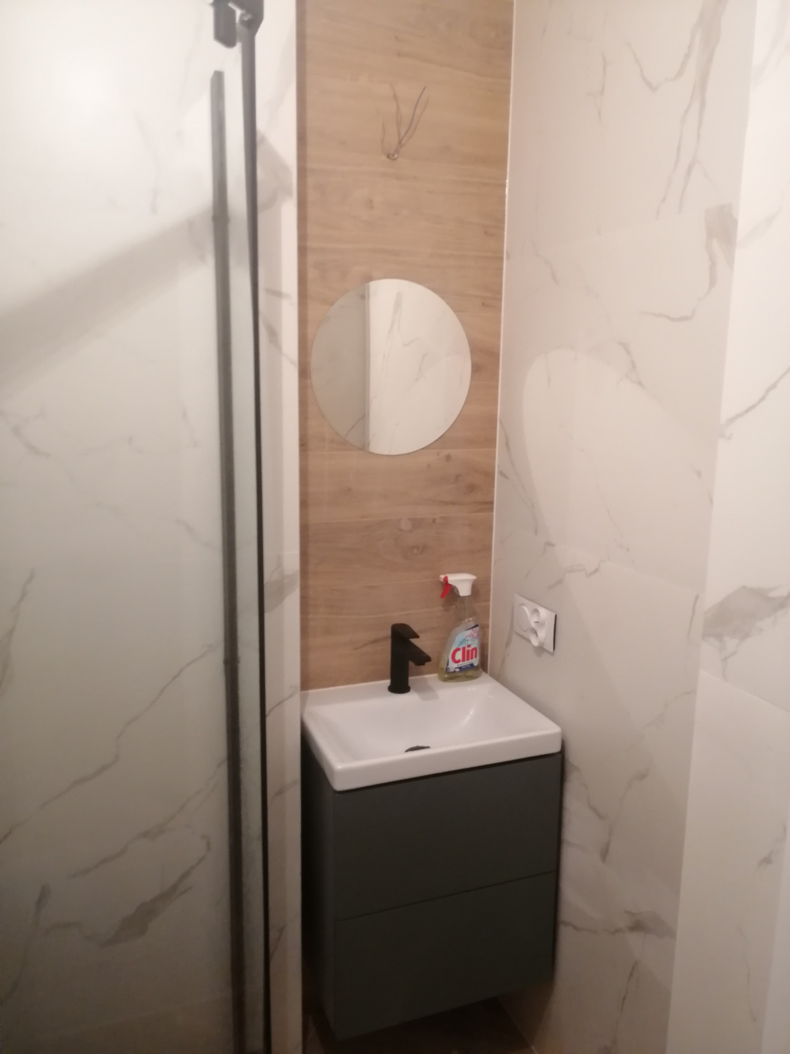 Nowoczesna łazienka po remoncie: umywalka z szafką w kolorze grafitowym, okrągłe lustro i płytki imitujące marmur na ścianach, widoczny fragment kabiny prysznicowej.