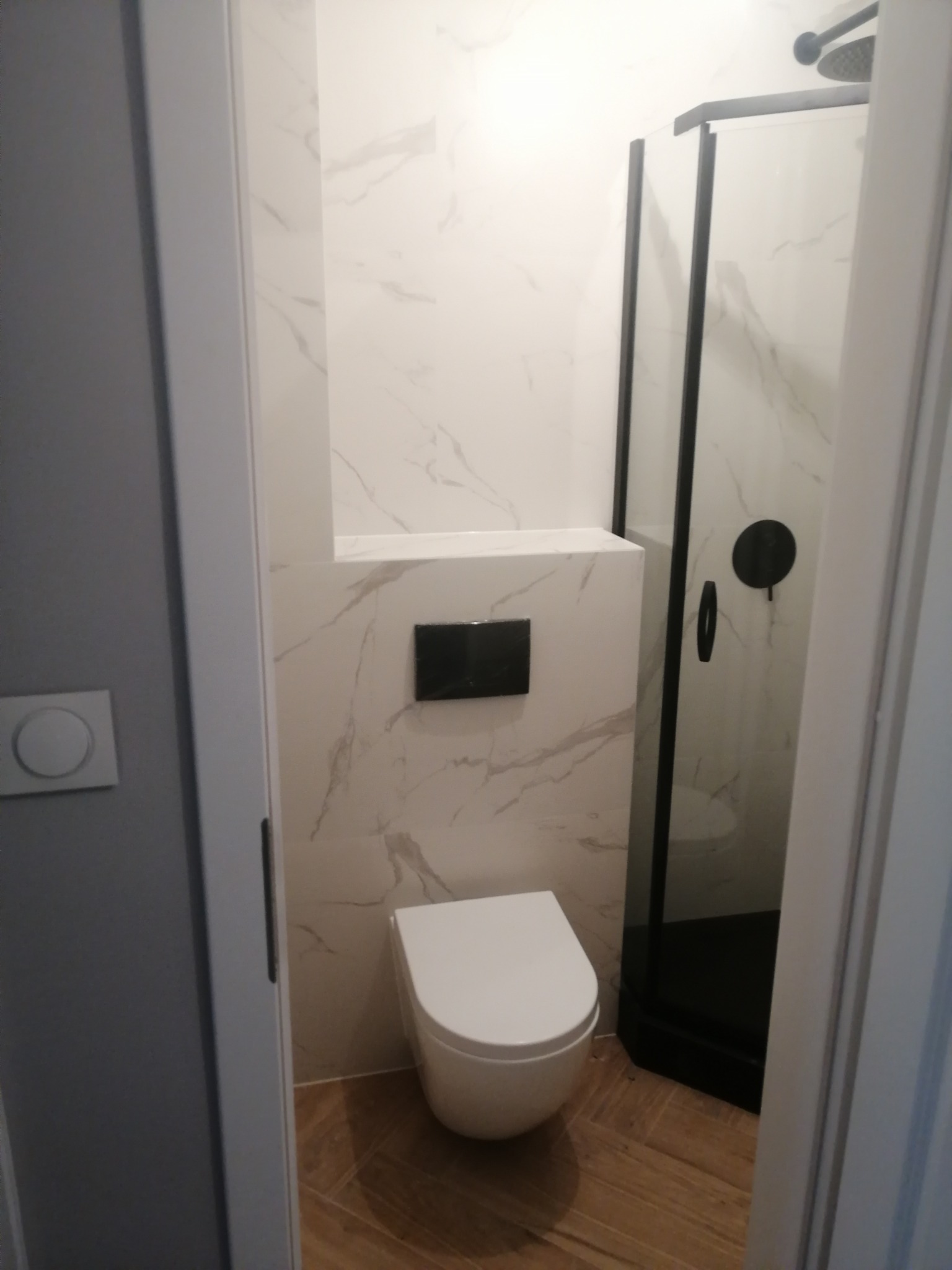 Nowoczesna łazienka z toaletą wiszącą, zabudowaną spłuczką, kabiną prysznicową z czarnymi profilami i białymi płytkami imitującymi marmur.