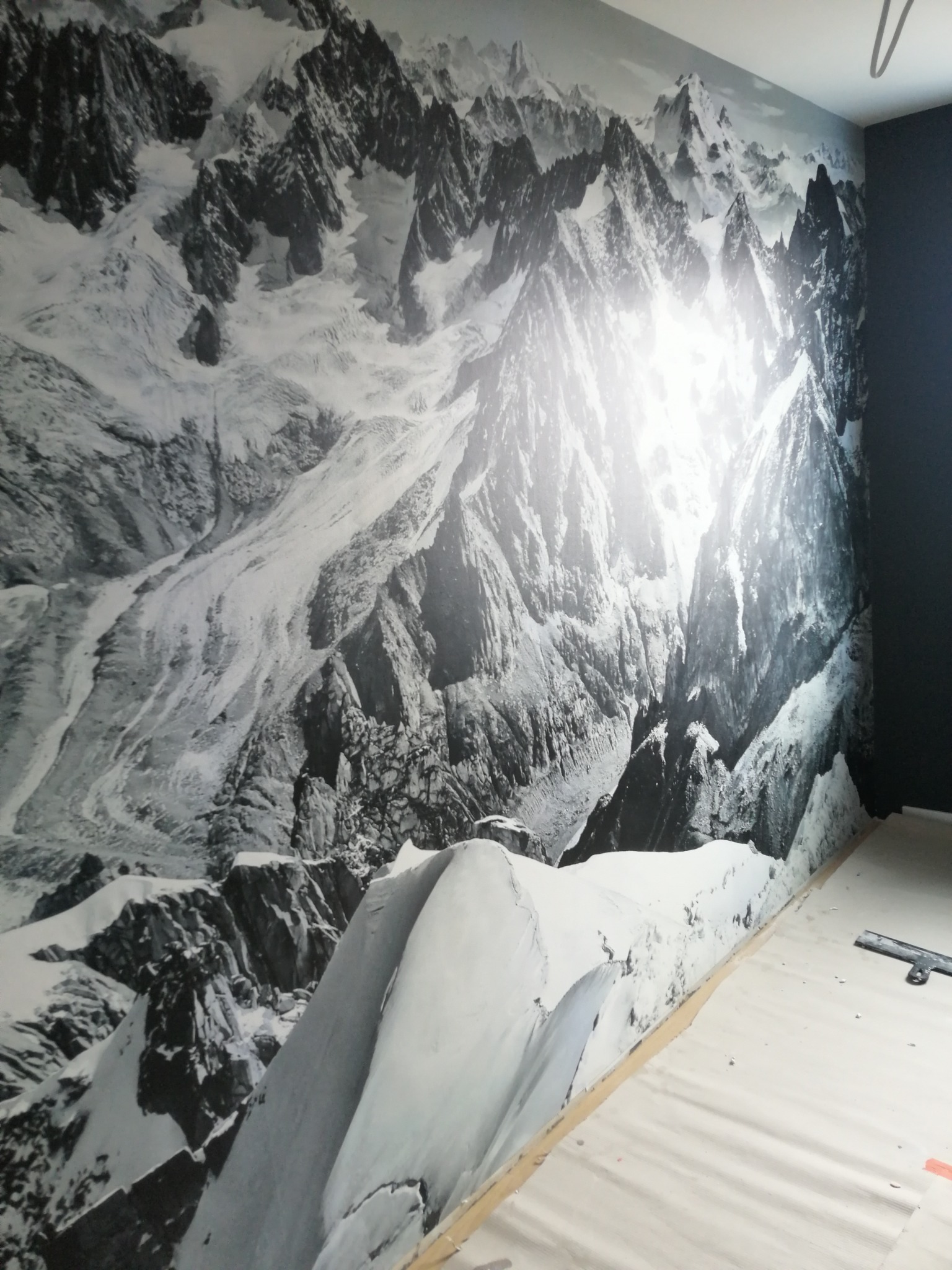 Fototapeta z motywem górskim w odcieniach szarości, częściowo przykryta folią ochronną na podłodze podczas montażu, widoczny fragment ściany pomalowanej na ciemny kolor.