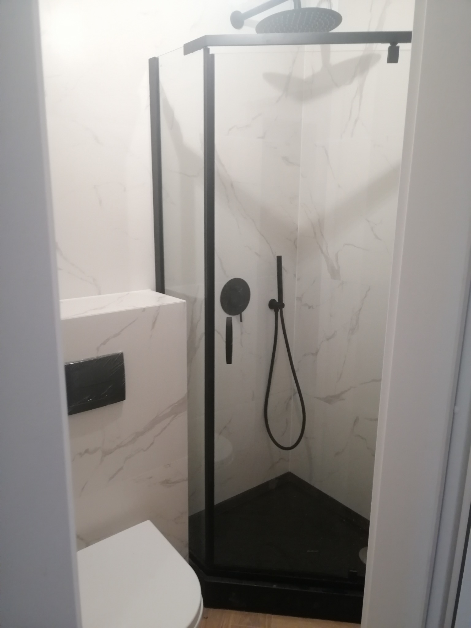 Nowoczesna kabina prysznicowa narożna z czarnymi profilami, deszczownicą i baterią, białe płytki imitujące marmur, wisząca toaleta z czarnym przyciskiem spłukiwania.