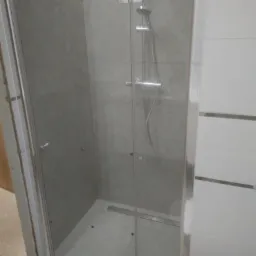 Szklana kabina prysznicowa z odpływem liniowym w podłodze, szare płytki na ścianach, chromowana armatura i uchwyty, białe płytki z poziomymi paskami na ścianie obok.