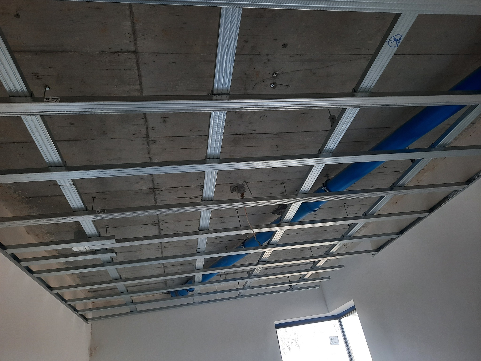 Stelaż metalowy podwieszany do sufitu z widocznymi niebieskimi rurami instalacyjnymi, przygotowany do montażu okładziny.